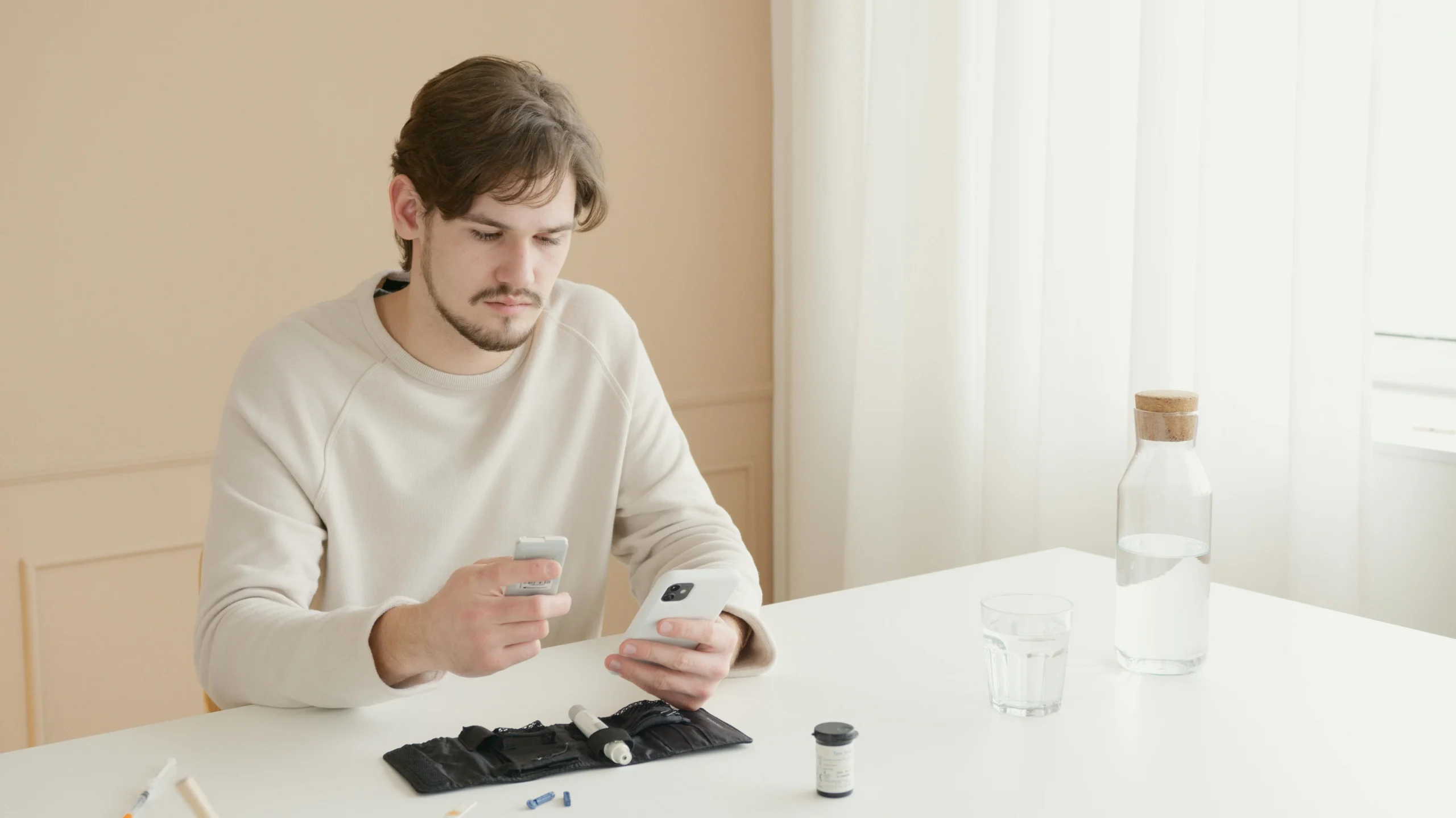 Man monitoring diabetes management with CGM and A1C tools, using a smartphone alongside diabetic equipment, water bottle, and glass in a bright minimalist setting.